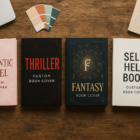 A selection of custom-designed book covers spread across a wooden desk, showcasing different genres like romance, thriller, fantasy, and self-help, with design tools and coffee in the background.